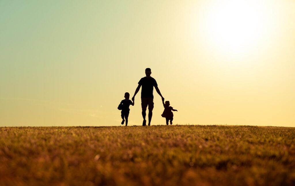 Dad walks hand-in-hand with two small children skipping alongside him as the sun begins to set