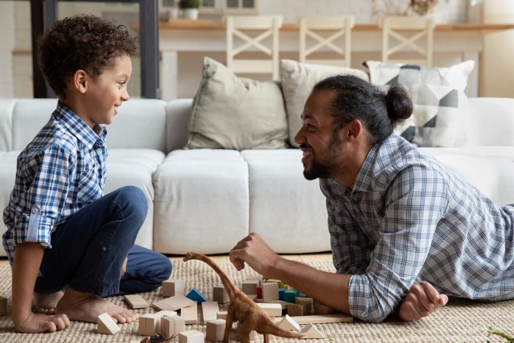 A boy and a young man - probably father and son - on the floor of their family room smiling at each other while playing with some wooden blocks and a miniature dinosaur.  